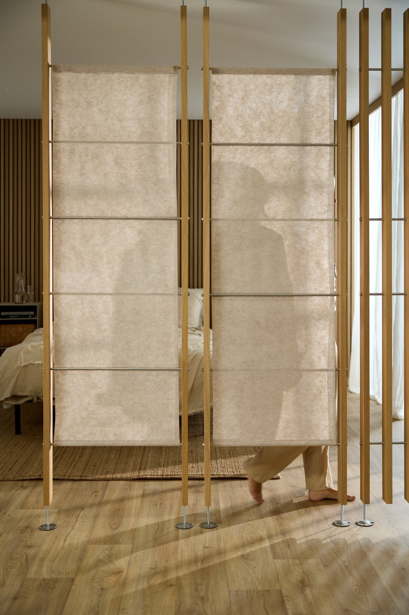 A person stands behind a WoodUpp Vigg Felt acoustic panel (58x176 cm, Light Beige) in a Nordic-style bedroom with wood floors and walls.